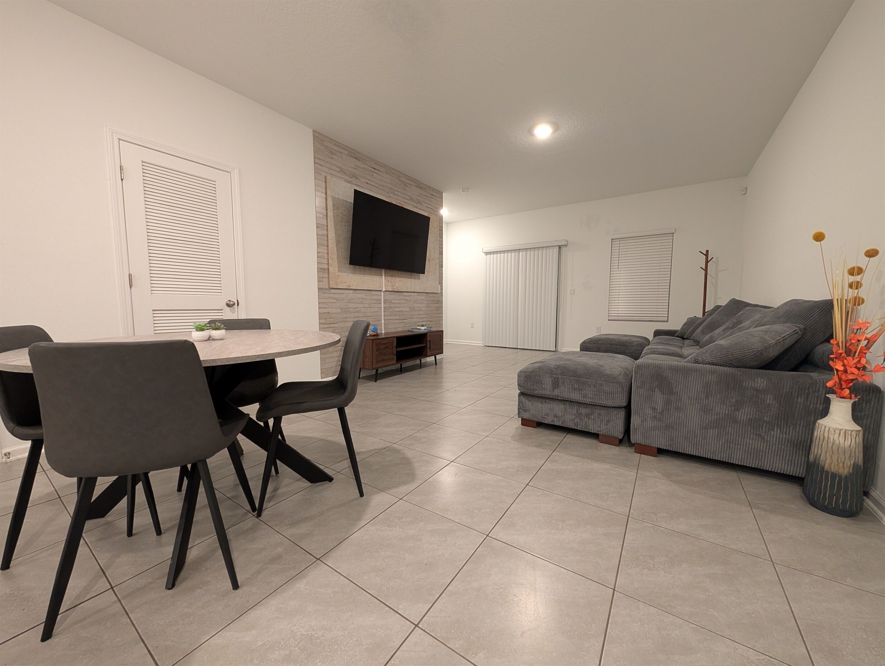 A modern living room with a grey sofa, a dining table with chairs, and a flat-screen TV mounted on the wall.