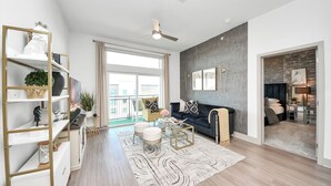 Ein modernes Wohnzimmer mit einem dunkelgrauen Sofa, einem Glastisch und einem weißen Teppich. Es gibt einen Deckenventilator, ein großes Fenster mit Vorhängen und einen Blick nach draußen.