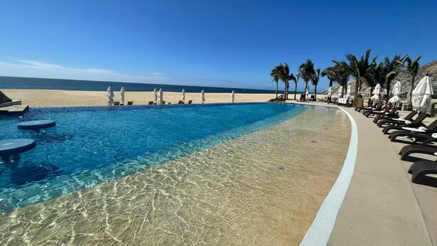 Ein Poolbereich mit klarem Wasser, einem Sandstrand und Liegestühlen.