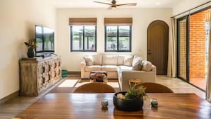 Ein modernes Wohnzimmer mit einer Couch, einem Couchtisch und einem Fernseher. Zwei Fenster mit Jalousien, ein Deckenventilator und eine Glastür, die zu einem Außenbereich führt.