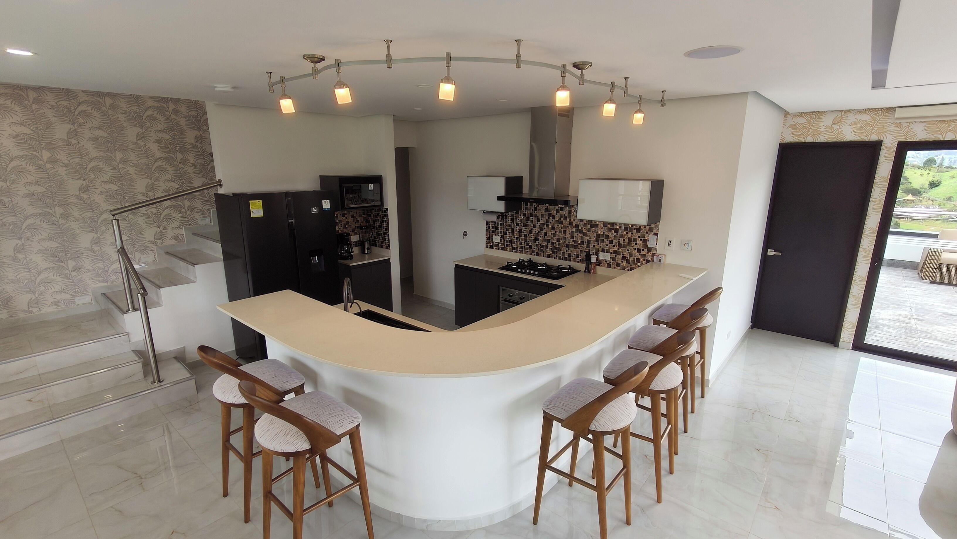 Una cocina moderna con una isla curvada, taburetes de bar y una escalera con barandilla de metal.