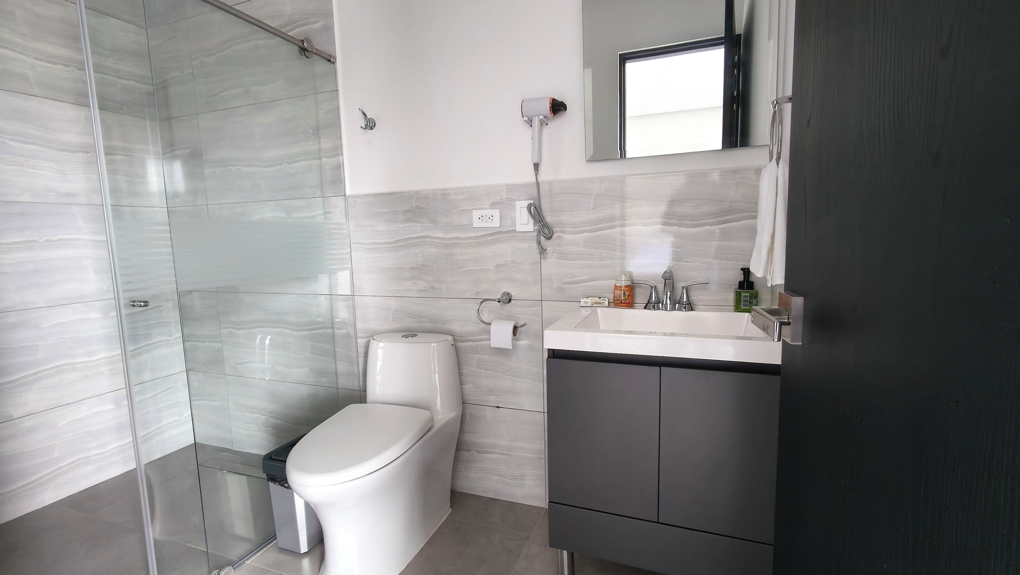 Un baño moderno con cabina de ducha de vidrio, inodoro, lavamanos con espejo y artículos de aseo.