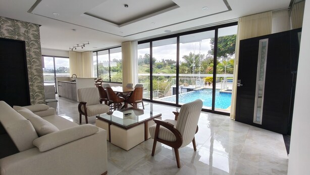 Un salón amplio con una mesa de centro de vidrio, asientos cómodos y un ventanal con vistas a una piscina y abundante vegetación.