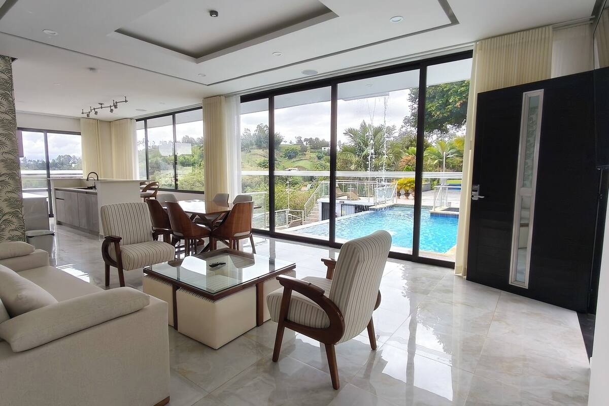 Un salón amplio con una mesa de centro de vidrio, asientos cómodos y un ventanal con vistas a una piscina y abundante vegetación.