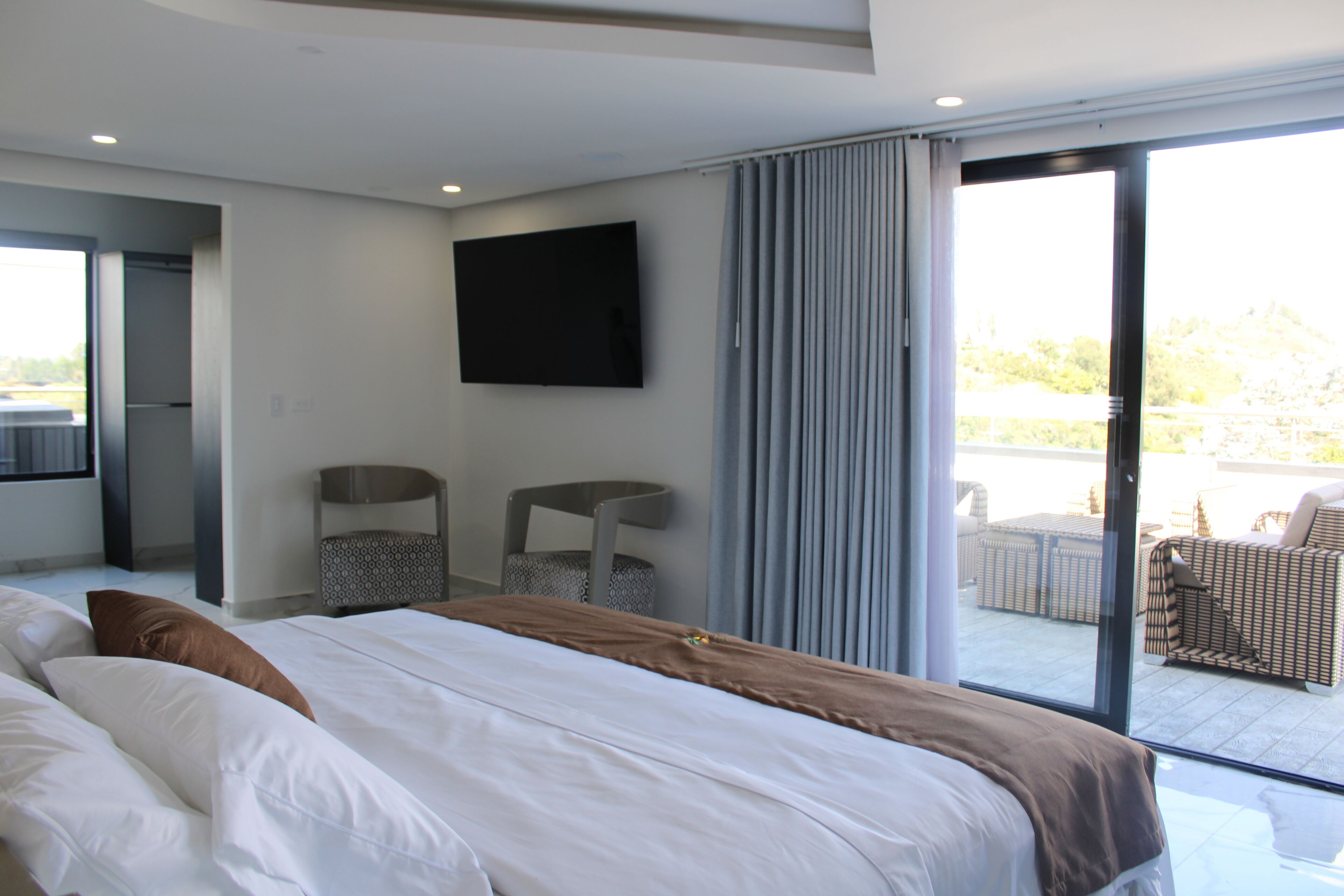 A modern hotel room with a large bed, a flat-screen TV mounted on the wall, and a balcony with outdoor furniture.
