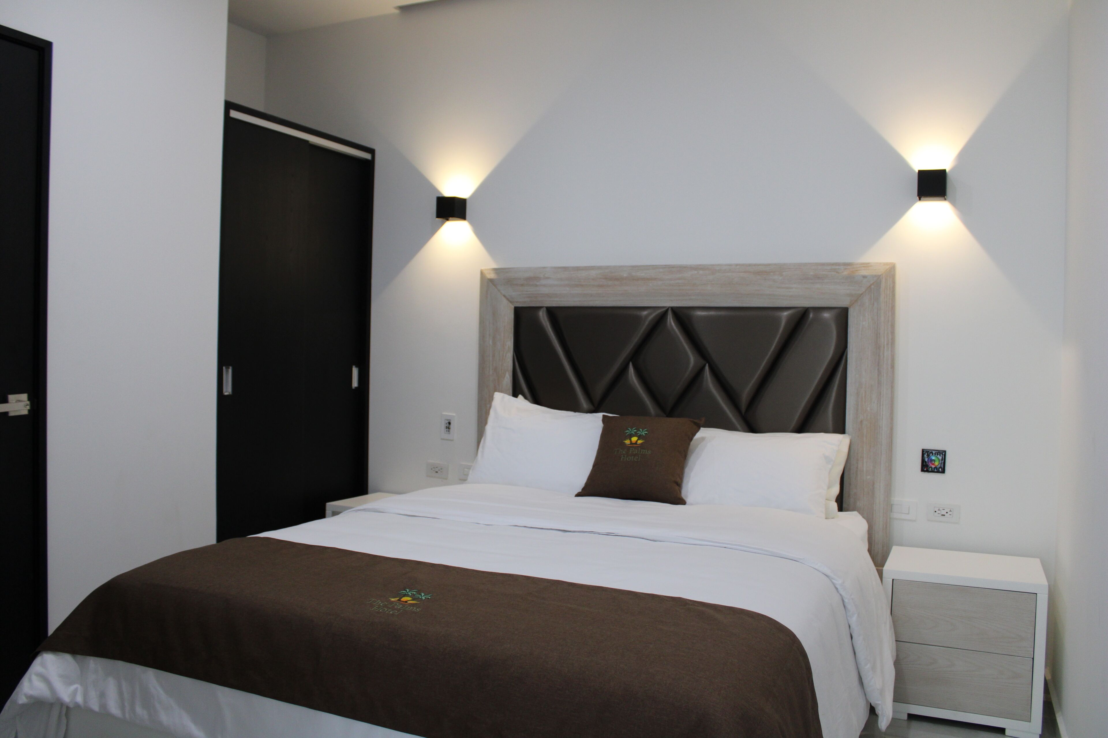 A neatly made bed with a wooden headboard, white bedding, and a brown throw blanket. There are two bedside tables with light-colored drawers and a wall-mounted light fixture.