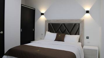 A neatly made bed with a wooden headboard, white bedding, and a brown throw blanket. There are two bedside tables with light-colored drawers and a wall-mounted light fixture.