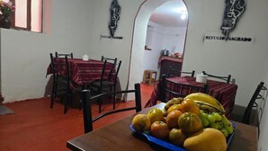 A dining area with tables and chairs, a fruit basket on the table, and a kitchen in the background. - REFUGIO SAGRADO BOUTIQUE (Ollantaytambo)
