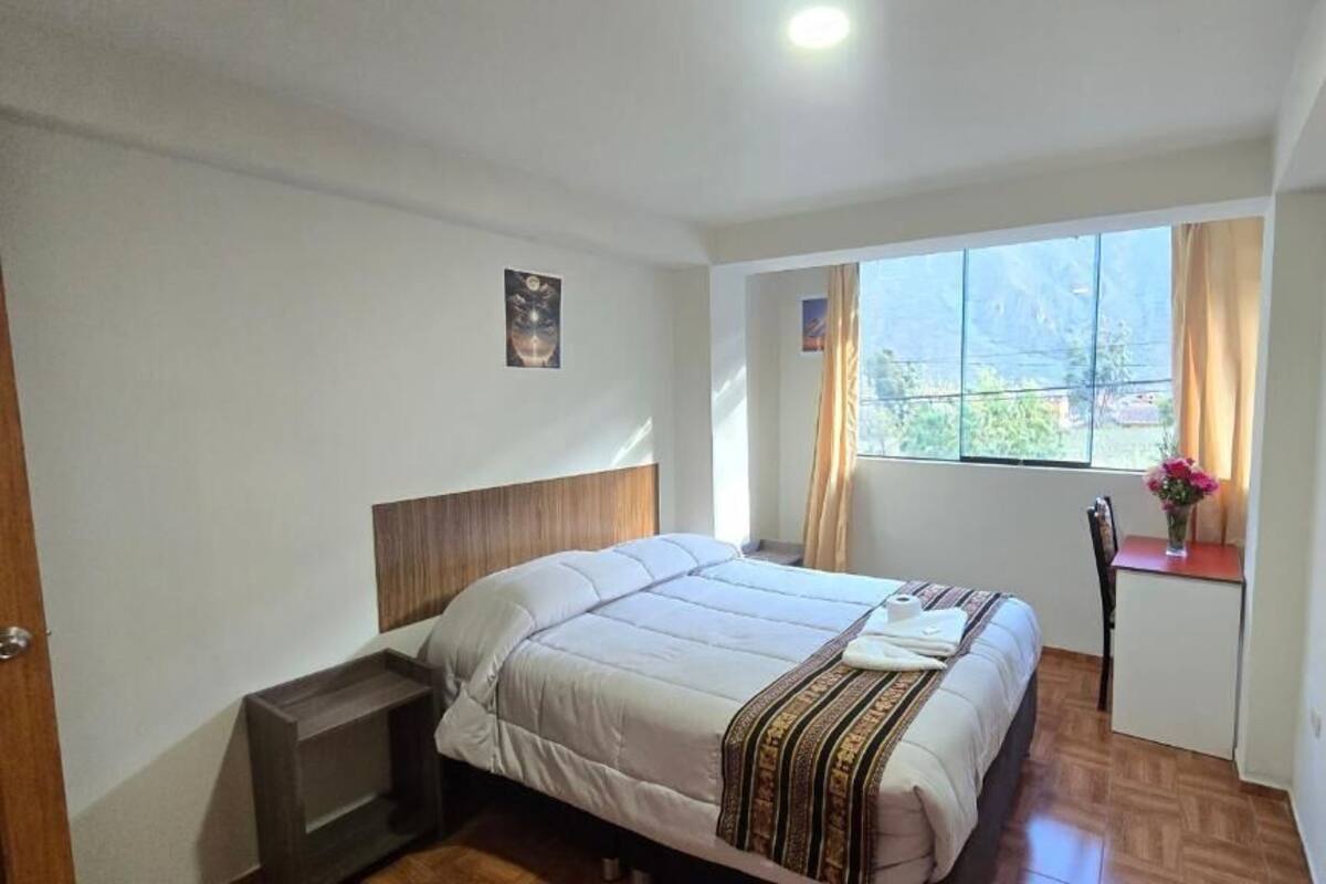 A bedroom with a bed, a small bedside table, a window with curtains, and a view of greenery outside.