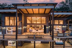 Una cabaña de madera de varios niveles con un balcón, ventanas amplias y un espacio central abierto con asientos.
