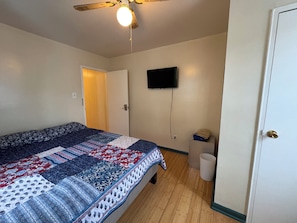 Een slaapkamer met een bed, een plafondventilator, een aan de muur bevestigde televisie, een deur en een afvalbak.