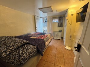 A bedroom with a bed, a bedside table, a window with curtains, a door, and a wall-mounted light fixture.