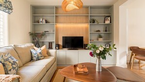 A modern living room with a sofa, coffee table, TV, and shelves with decorative items. - Your Place - 2 bedroom Mews House in Notting Hill (London)