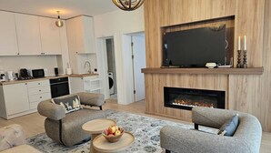 En moderne stue med pejs, tv og et køkkenområde med hvide skabe.