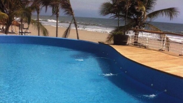 Une piscine d’un bleu éclatant, bordée de carreaux et dotée d’un escalier en métal, qui offre une vue sur une plage parsemée de palmiers et sur l’océan.