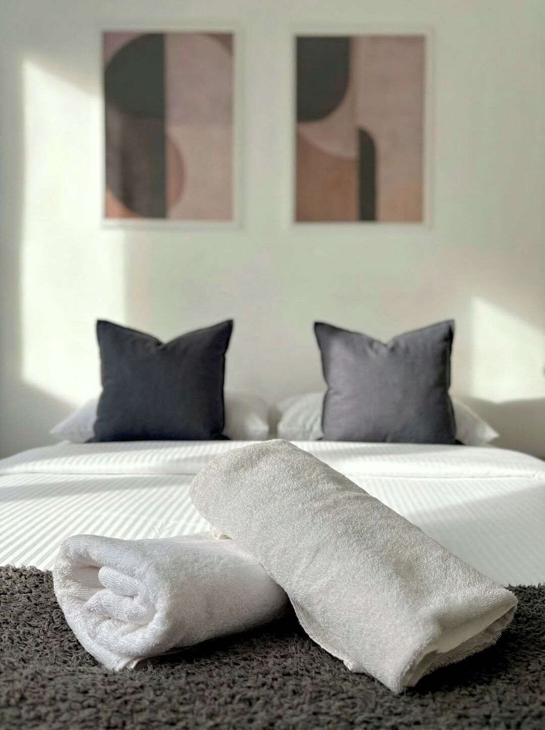 A neatly made bed with a white duvet, two grey pillows, and a rolled white towel on a grey carpet.