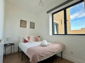 Une chambre à coucher avec un lit, une petite table, une fenêtre donnant sur un immeuble en briques et des œuvres d’art abstraites accrochées au mur.