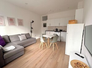 Un apartament modern amb un sofà gris, una taula de dinar amb cadires, una rentadora i un televisor.