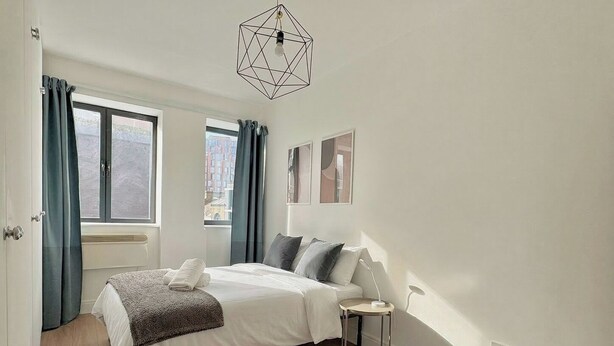 A bedroom with a bed, a bedside table, a geometric light fixture, and a window with curtains. - Shoreditch Apartments by PrimeCityHosts F6 (Greater London)