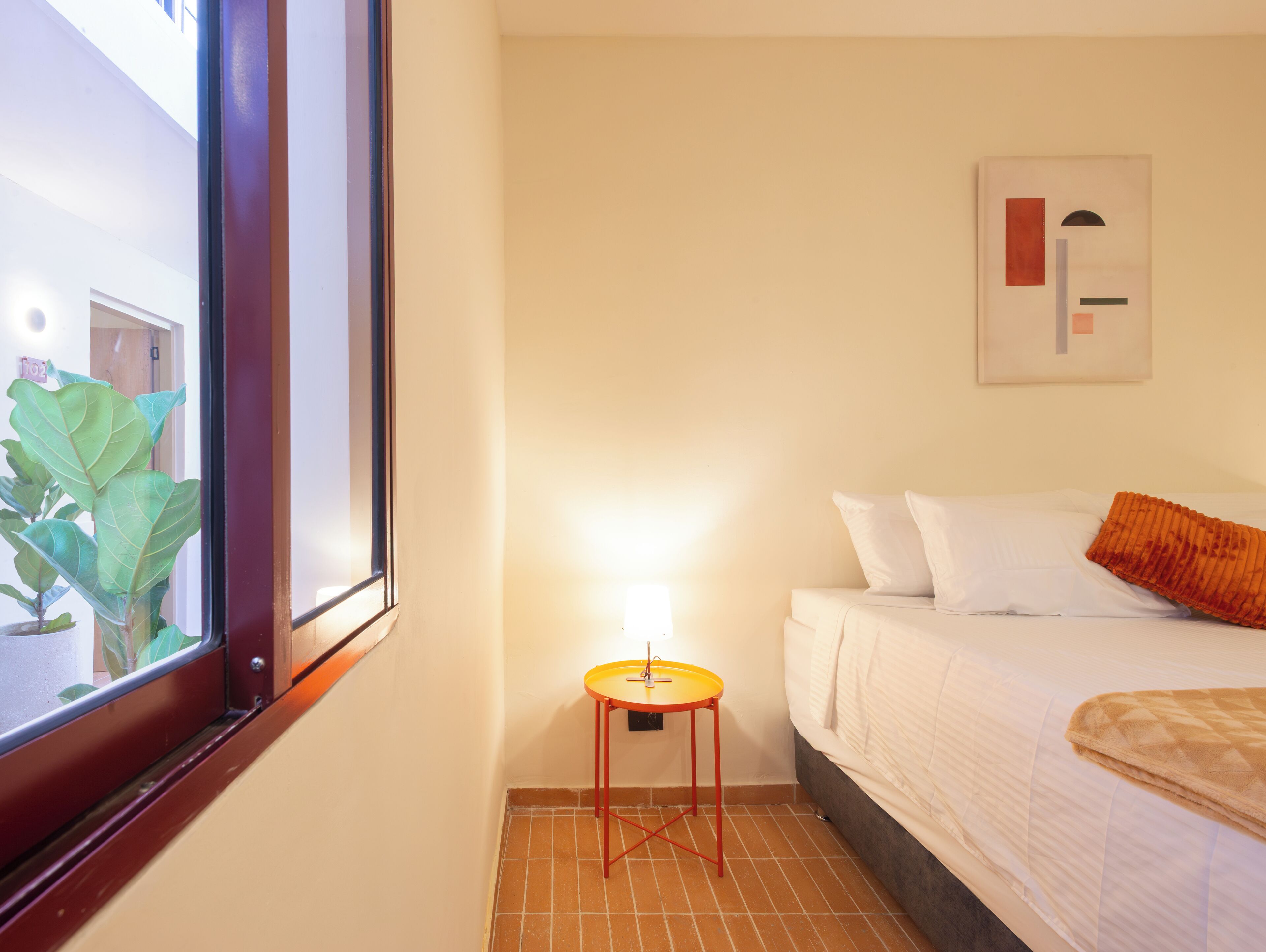 A hotel room with a bed, a small table with a lamp, a window with a view of a building, and a plant outside.