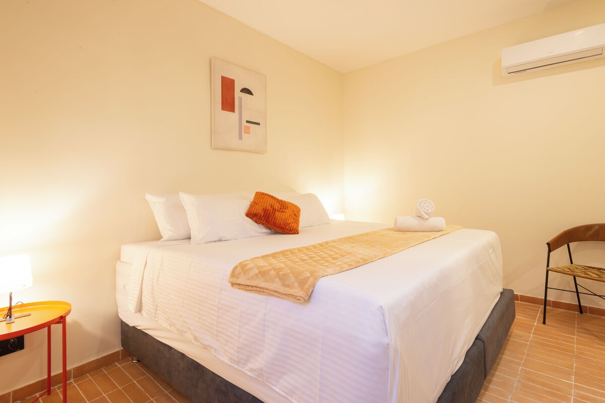 Una cama bien hecha con sábanas blancas y una manta beige, una silla de madera, una mesita con base roja y un equipo de aire acondicionado en la pared.