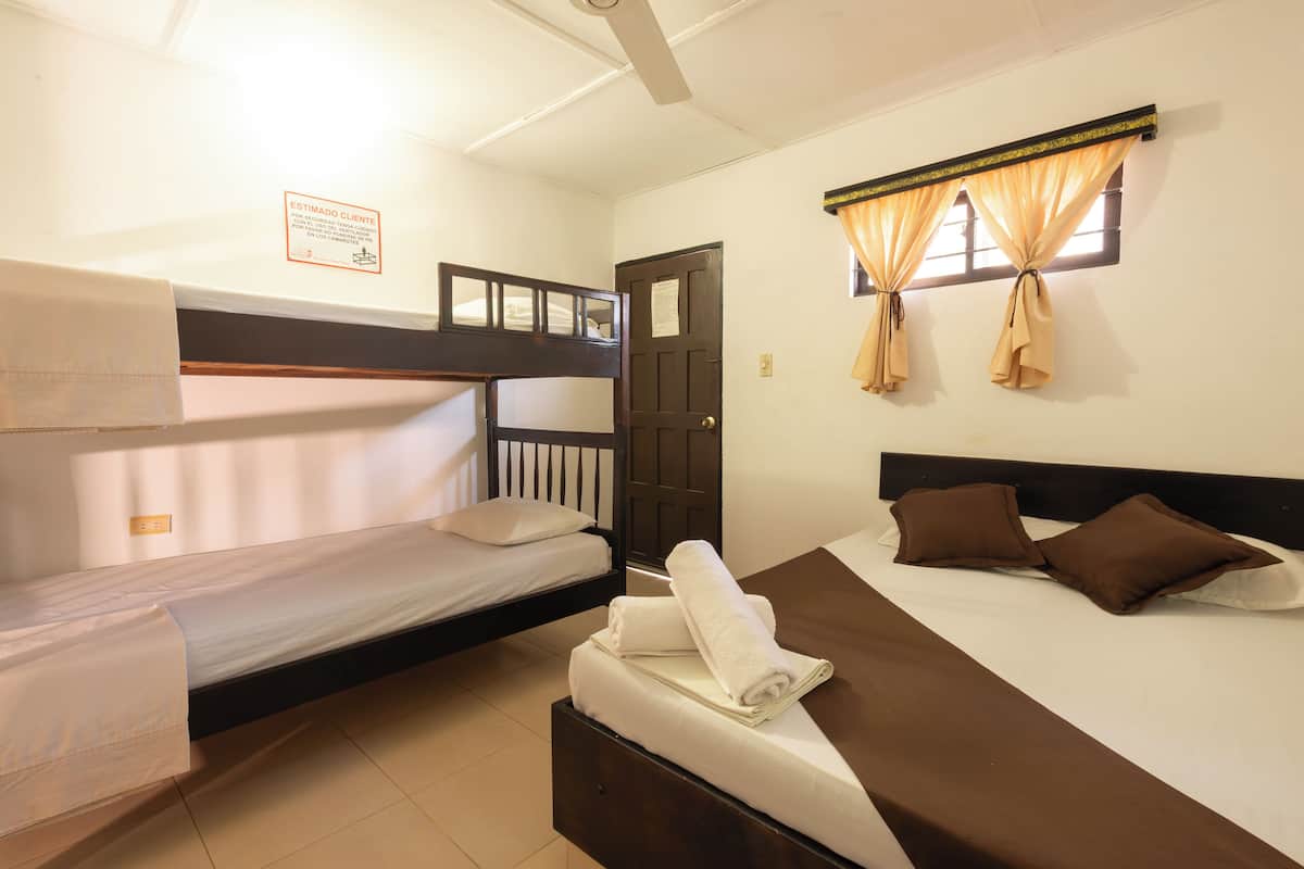 A room with two beds, one with a bunk bed and the other with a single bed. There is a door, a window with curtains, and a sign on the wall.