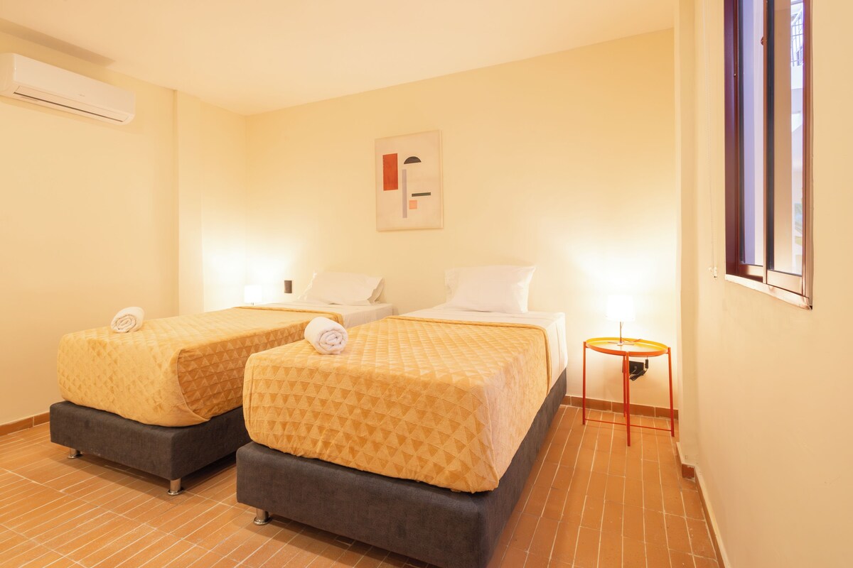 Habitación de hotel con dos camas, una mesita, una lámpara y un cuadro en la pared.
