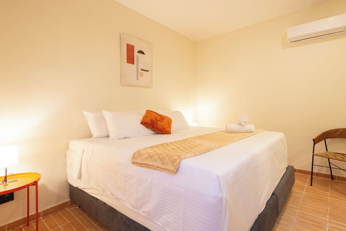 A neatly made bed with white linens and a beige throw blanket, a wooden chair, a small table with a red base, and an air conditioning unit on the wall.