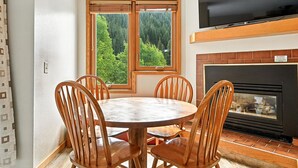 A dining area with a round table and four wooden chairs, a large window with a view of a forest, a flat-screen TV mounted above the fireplace, and a brick fireplace. - Ski in/ski out condo at Winter Park - cozy slopeside with pool, hot tub & more (Winter Park)