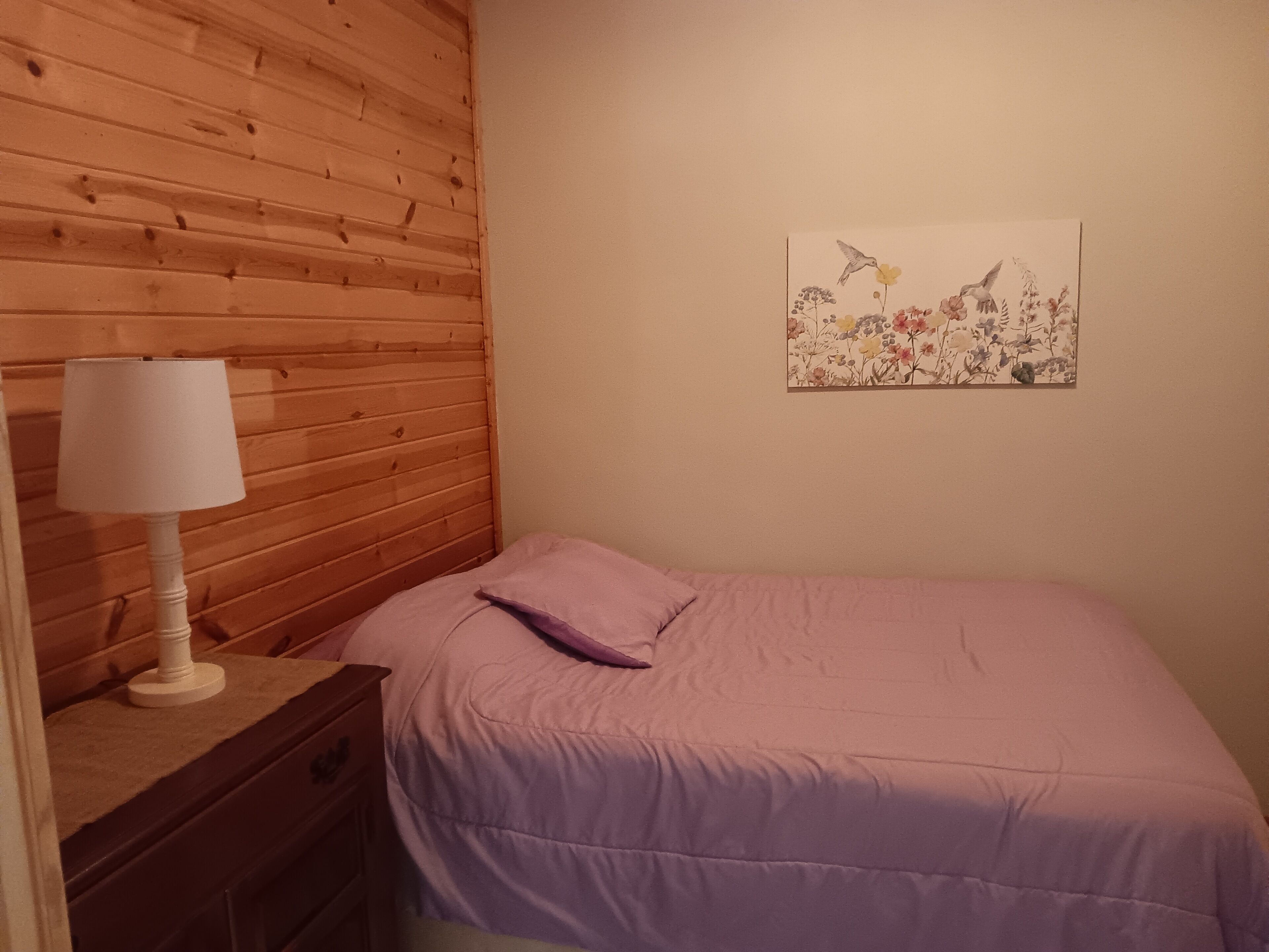 Un dormitorio con cabecera de madera, una mesita de noche con lámpara, cama con colcha morada y una obra de arte floral en la pared.