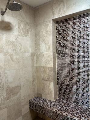 Ein Badezimmer mit beigen Marmorfliesen, einer Dusche mit Lochduschkopf und einer mosaikverzierten Sitzbank.