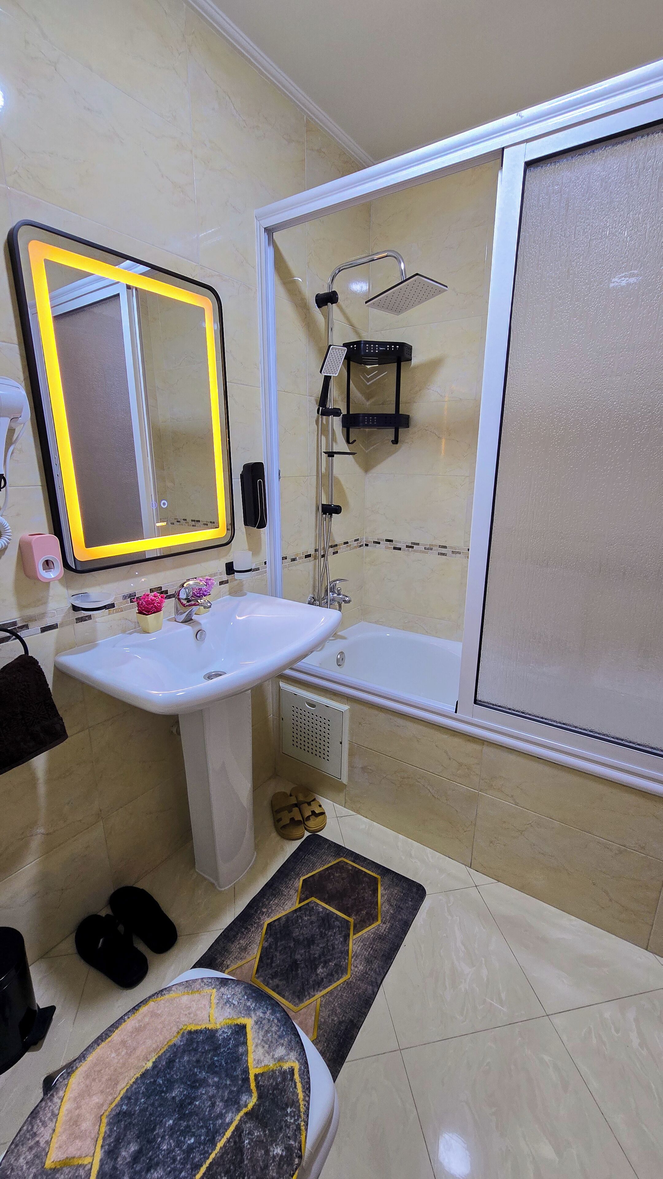 Un baño con lavamanos blanco, espejo y ducha. Hay un perchero y un estante pequeño con artículos de aseo.