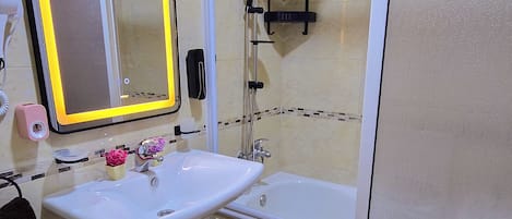 Un baño con lavamanos blanco, espejo y ducha. Hay un perchero y un estante pequeño con artículos de aseo.