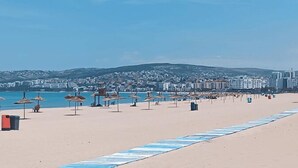 Een strand met parasols en stoelen, een heldere lucht en gebouwen op de achtergrond.