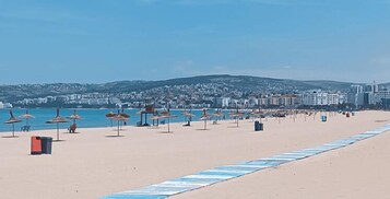 A beach with umbrellas and chairs, a clear sky, and buildings in the background.