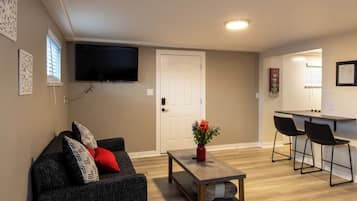 Un salon comprenant un canapé, une table basse et un téléviseur fixé au mur. On y trouve une porte donnant sur une autre pièce, ainsi qu’une petite table avec des chaises.
