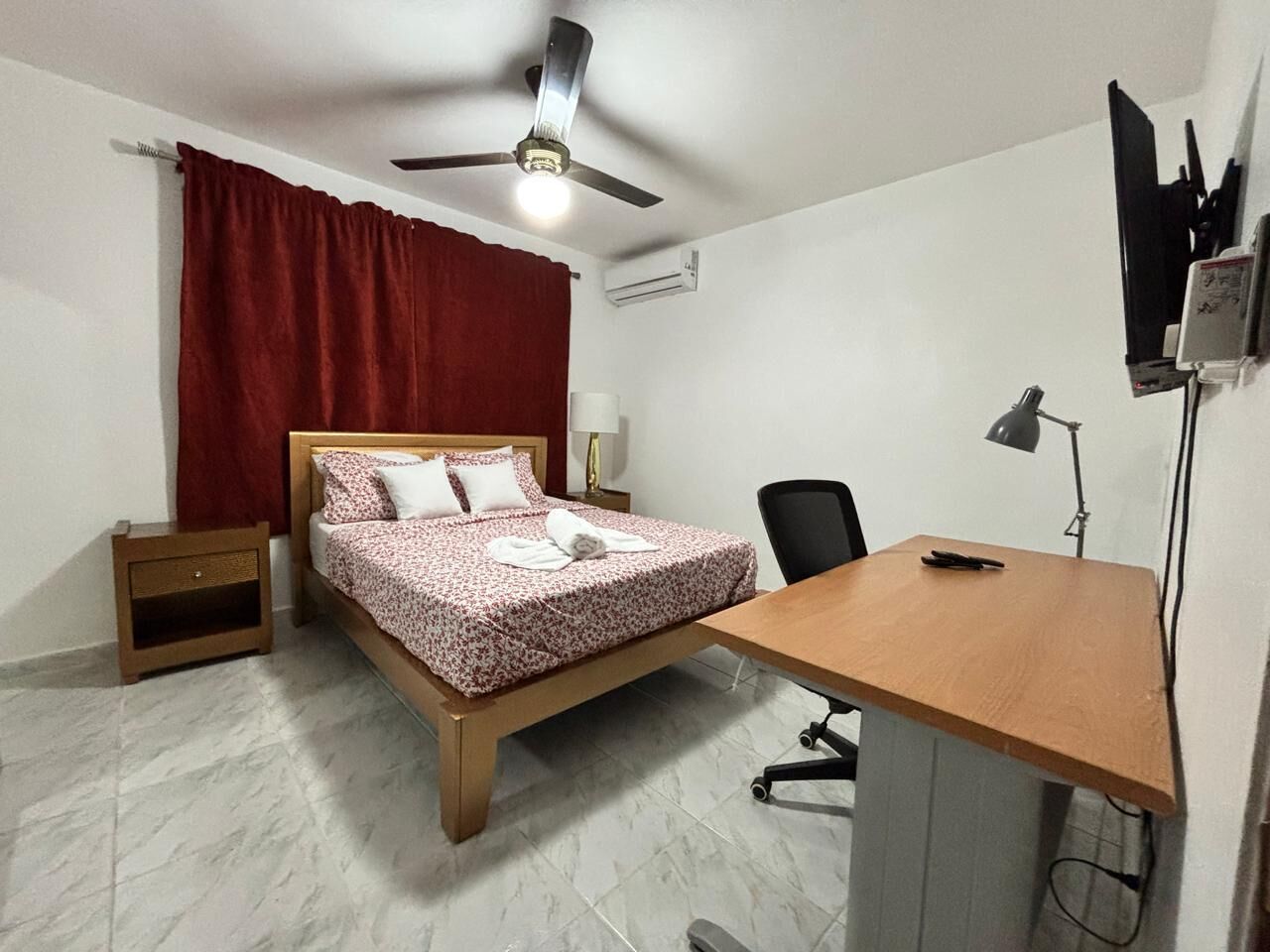 Une chambre à coucher comprenant un lit, un bureau, une chaise, un ventilateur de plafond, un climatiseur et un téléviseur fixé au mur.
