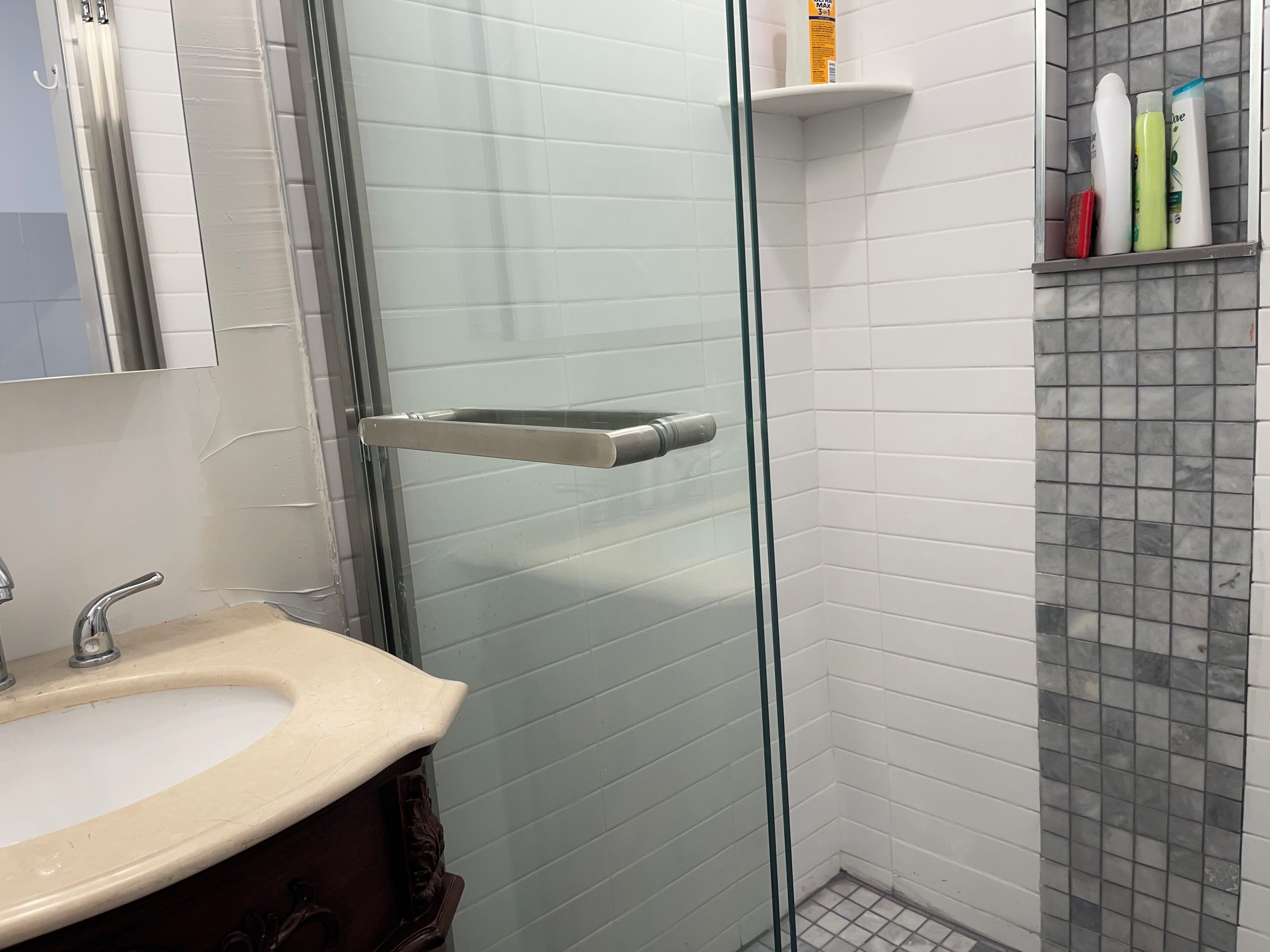 Et badeværelse med glasbrugedør, et beige håndvask og toiletartikler på en hylde.