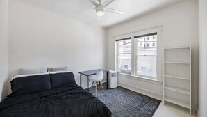 A bedroom with a bed, a desk, a chair, a ceiling fan, and a window with a view of buildings. - Stylish Downtown LB Flat – Walk to Everything! (Long Beach)