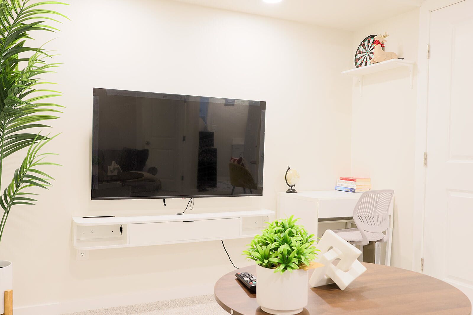 Un salon avec un téléviseur fixé au mur, une plante en pot, une télécommande et une chaise blanche.