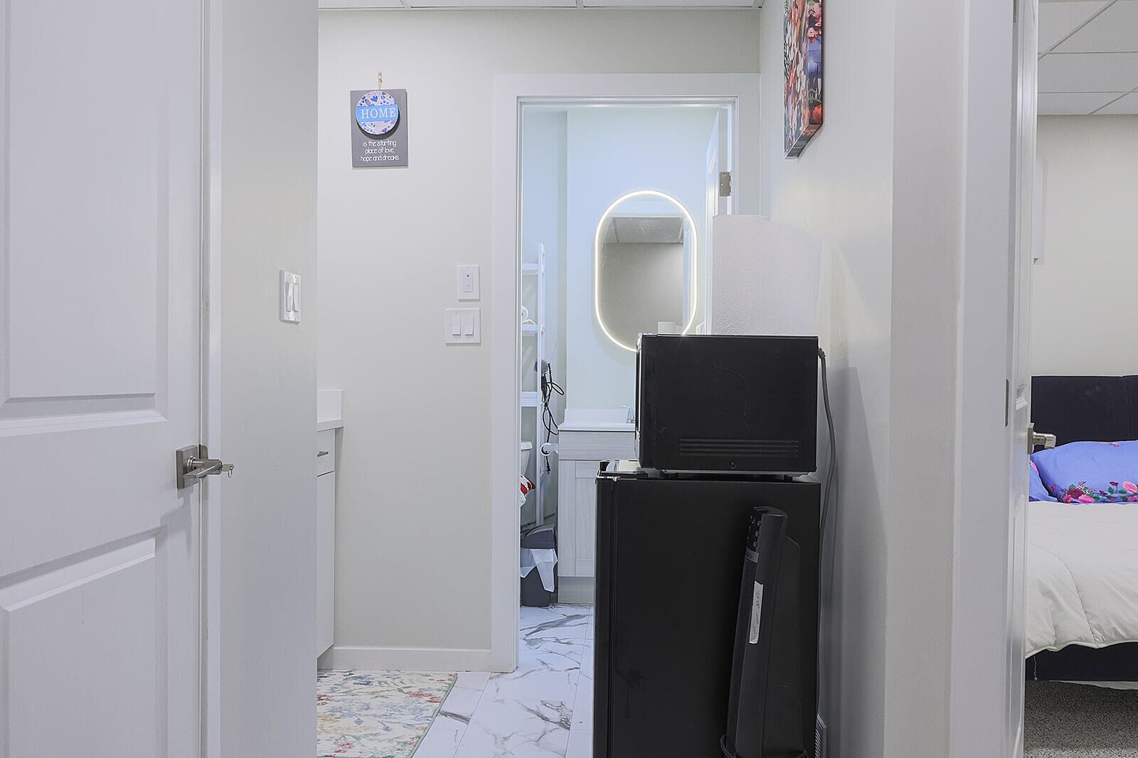 A narrow hallway with a door leading to a bathroom, a bed with a floral bedspread, and a black refrigerator.