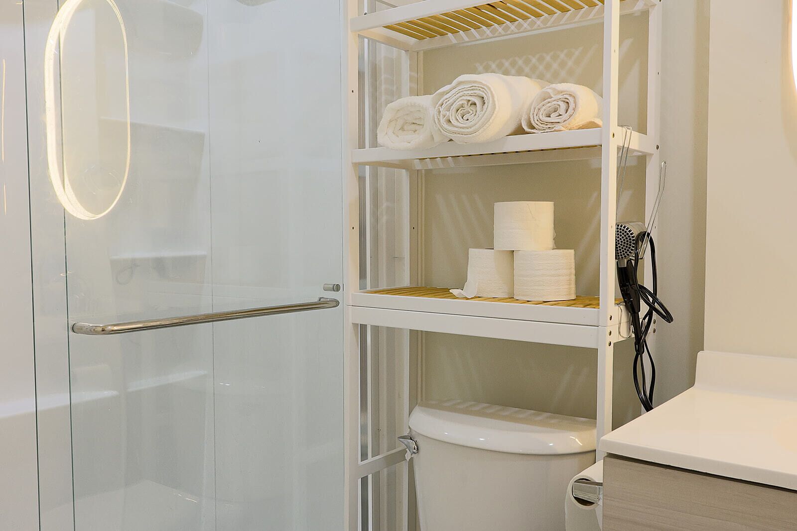Un baño con cabina de ducha de vidrio, inodoro y repisa con toallas y papel higiénico.