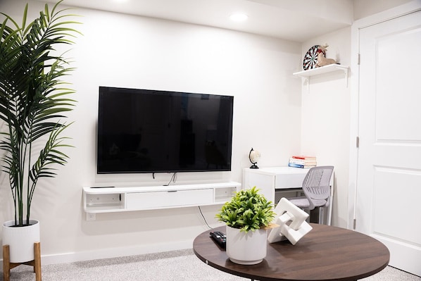 Una sala de estar con televisor de pantalla plana montado en la pared, una consola blanca, una mesa de centro redonda de madera y una planta en maceta.