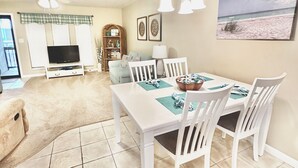Uma área de refeições com uma mesa e cadeiras brancas, um ventilador de teto e uma pintura de uma paisagem praiana.