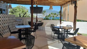 An outdoor dining area with wooden tables and black chairs, a thatched roof, and a view of the sea and mountains. - Beautiful apartment in Residencial Astorias! (Acapulco de Juárez)