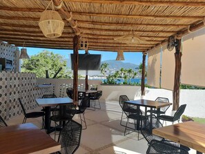 An outdoor dining area with wooden tables and black chairs, a thatched roof, and a view of the sea and mountains.