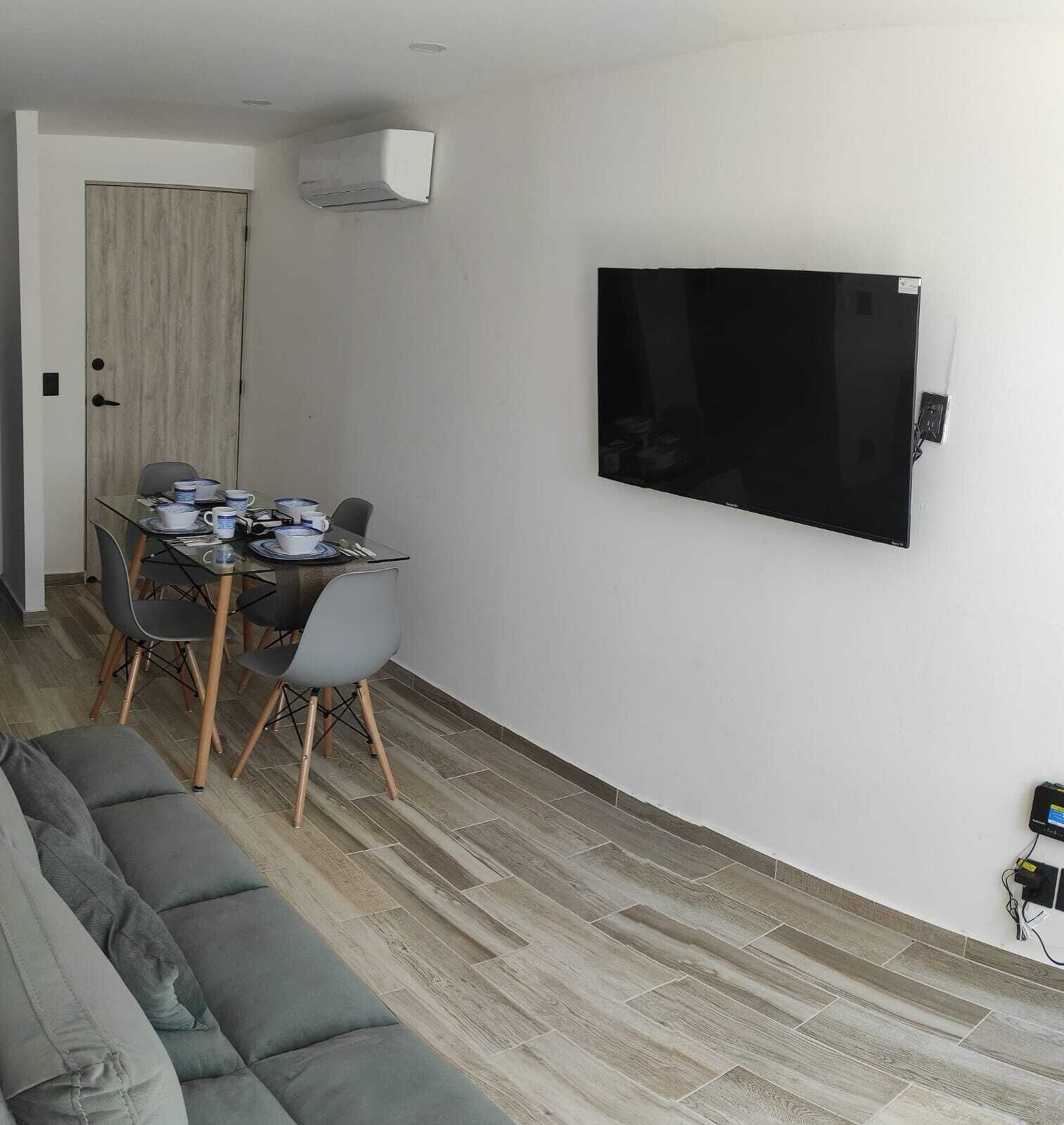 Un salon comprenant un canapé, une table à manger avec des chaises, un téléviseur à écran plat fixé au mur et une porte avec un panneau en bois.