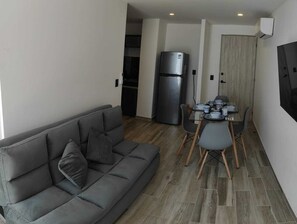A compact living space with a grey sofa, a dining table with chairs, a refrigerator, and a wooden door.
