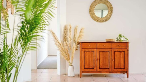 Een houten kast met een spiegel erboven, een witte vaas met pluimgras, en een plant op de voorgrond.