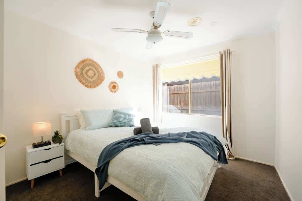 Un dormitorio con cama, mesita de noche, ventilador de techo y ventana con cortinas.